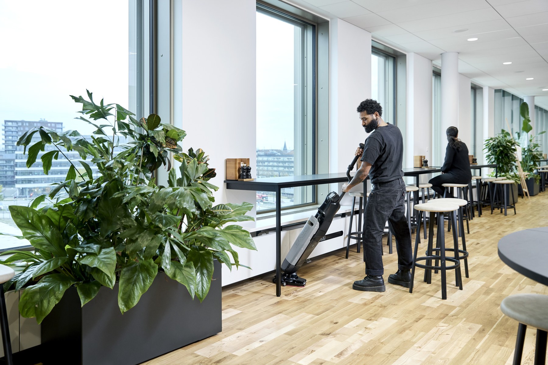 Cleaning operative using a Nilfisk Dryft compact scrubber dryer on a wood floor in a modern corporate canteen with floor-to-ceiling windows and a city skyline beyond