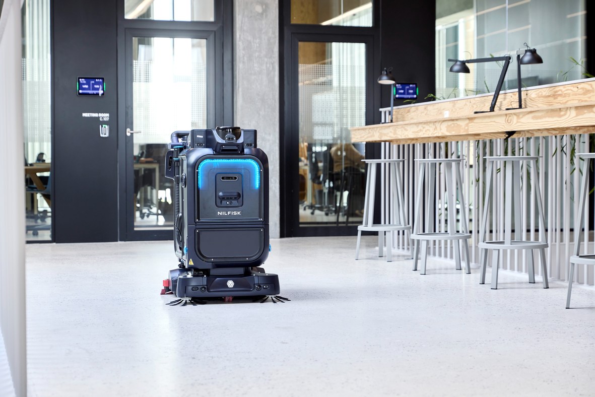 Nilfisk SC25 autonomous robotic scrubber dryer cleaning a polished concrete floor outside a glass-walled meeting room in a modern corporate office