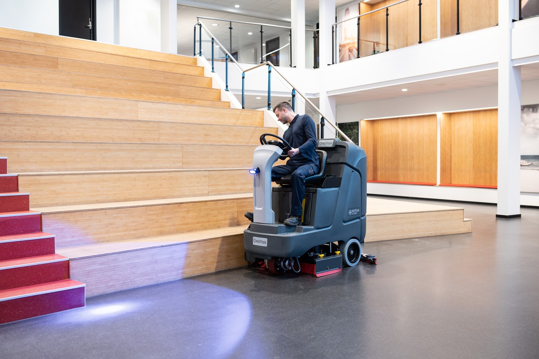 Operator on a Nilfisk SC4000 ride-on scrubber dryer cleaning the dark vinyl floor of a modern college atrium, blue UV floor projection visible alongside bamboo tiered seating