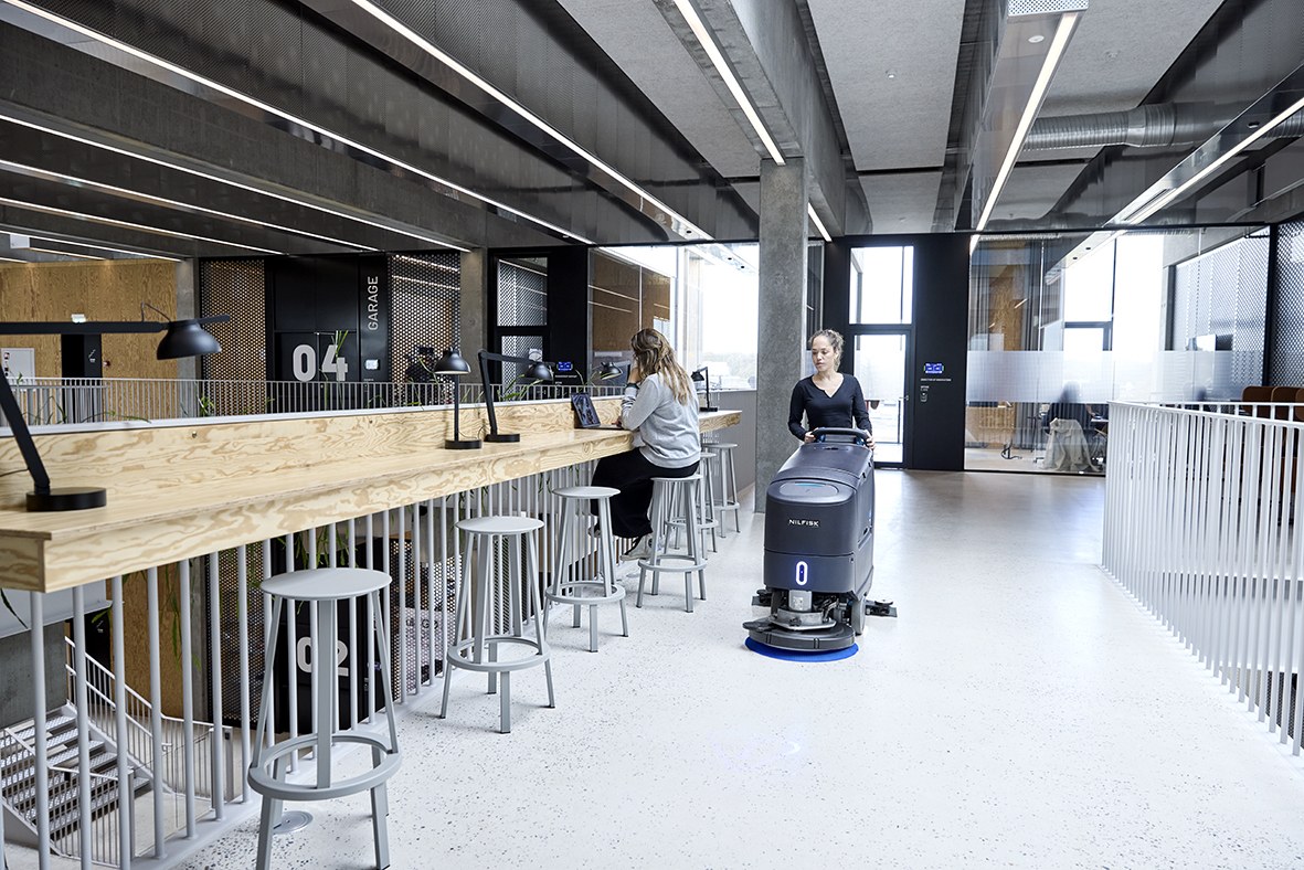 Female operator with a Nilfisk SC550 walk-behind scrubber dryer cleaning the polished concrete floor of a college study space, students working at laptops at the bar-height counter alongside