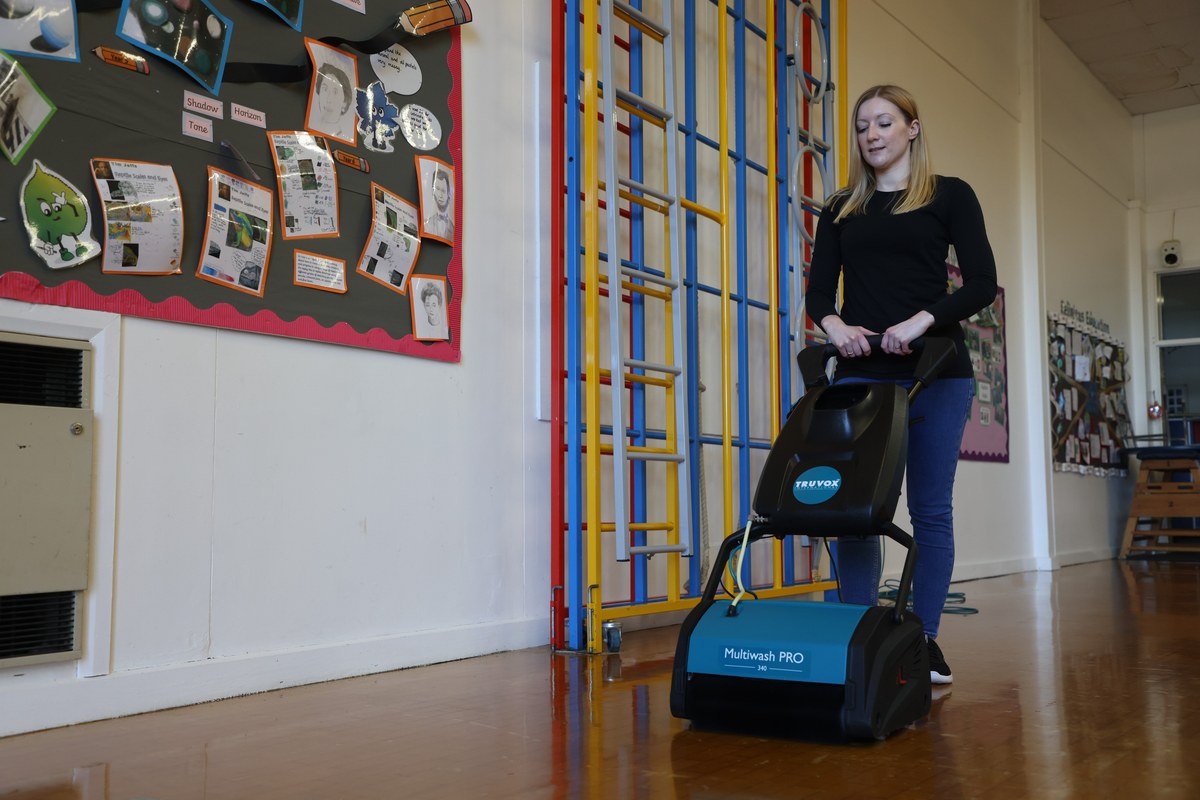 Operator using a Truvox Multiwash PRO 340 on the polished wooden floor of a primary school hall, climbing frame and pupil artwork visible behind