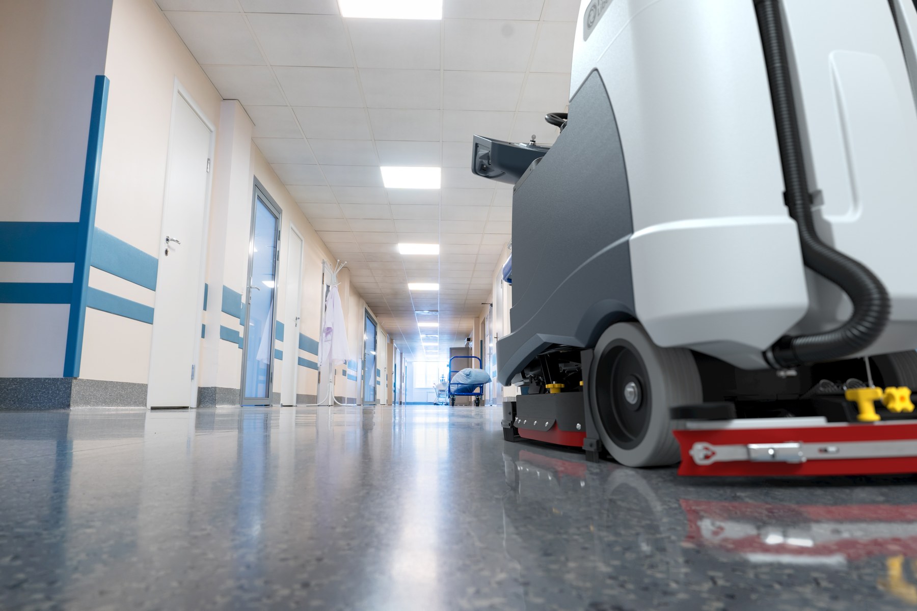 Low-angle view down a hospital corridor with a Truvox walk-behind scrubber dryer cleaning the polished vinyl floor in the foreground