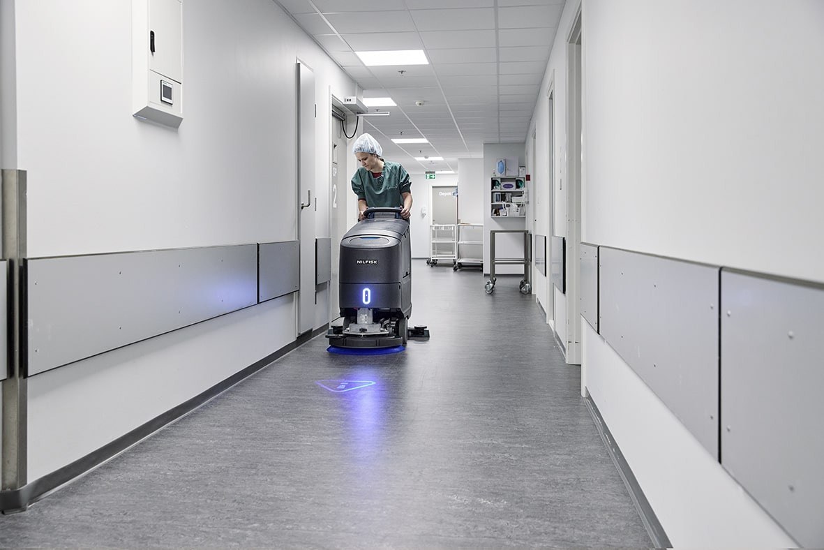 Operator in green scrubs and hairnet pushing a Nilfisk scrubber dryer down a clinical corridor with UV floor projection