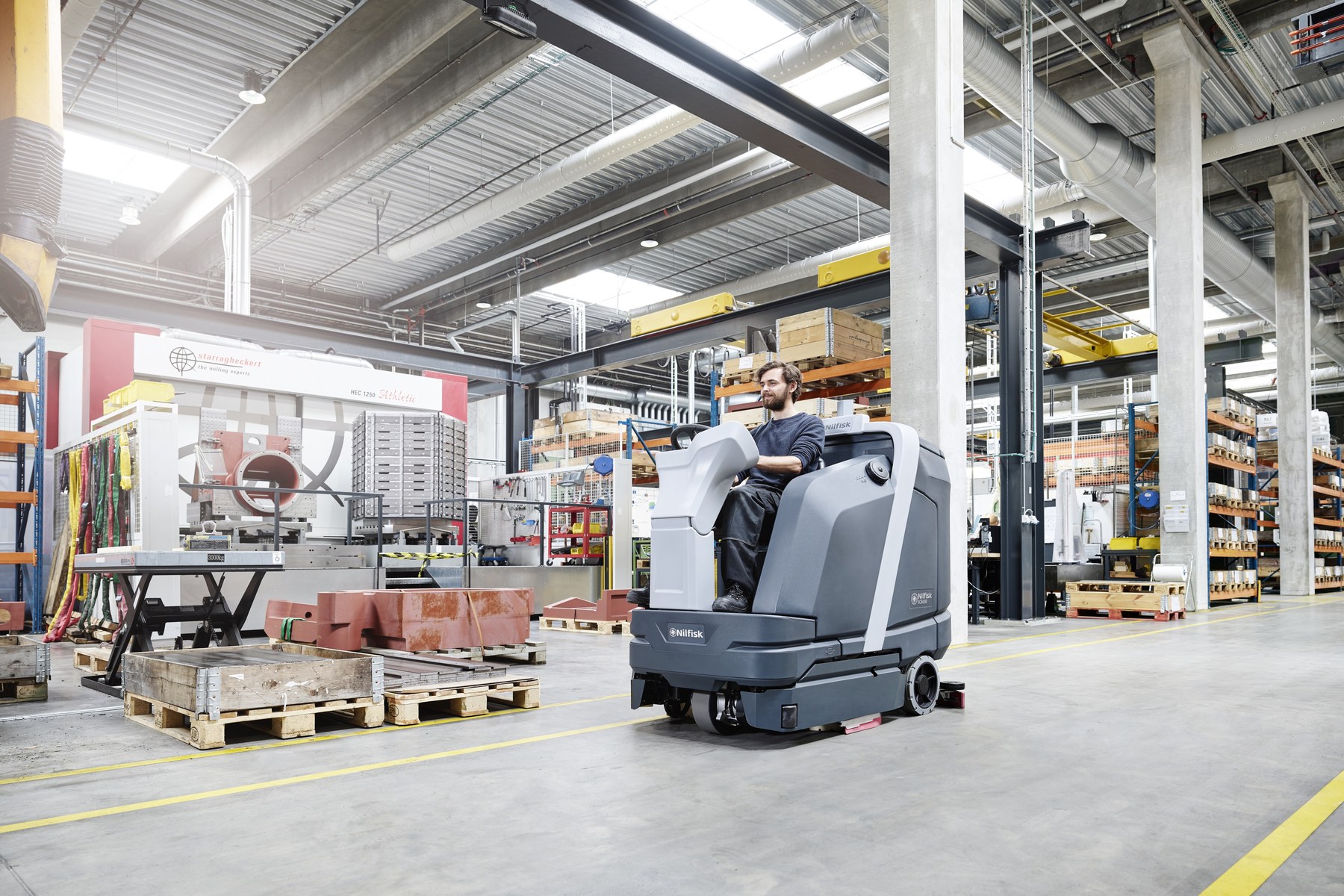 Operator on a Nilfisk SC6000 ride-on scrubber dryer cleaning the polished concrete floor of an engineering production facility, milling machinery and palletised stock either side