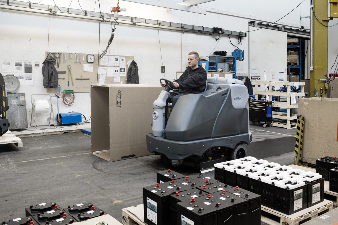 Operator on a Nilfisk SC6500 ride-on scrubber dryer manoeuvring around battery banks and packaging in an industrial service workshop