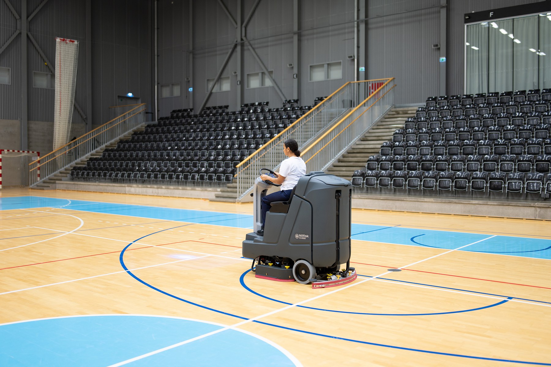 Operator on a Nilfisk SC4000 ride-on scrubber dryer cleaning the sprung wood floor of a multi-court sports arena, empty tiered seating in the background