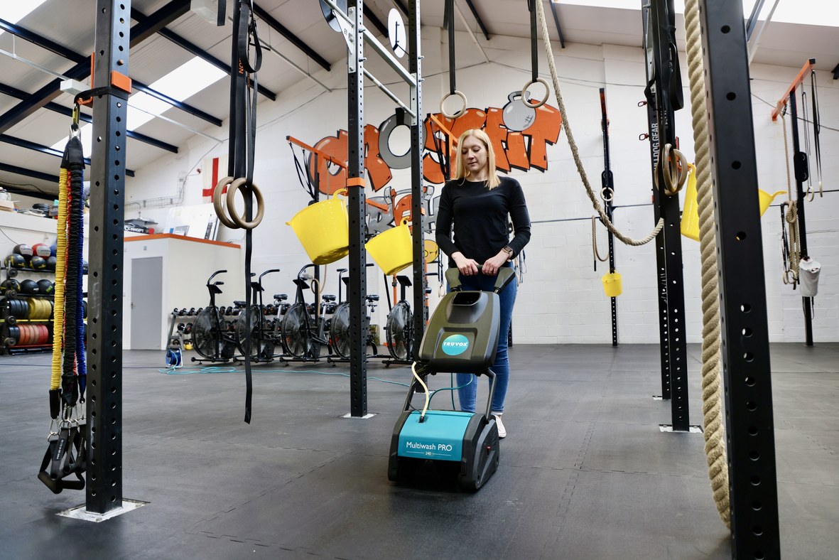 Female operator with a Truvox Multiwash PRO 240 cleaning the rubber matting of a CrossFit gym, gymnastic rings and rope climbs surrounding the cleaning area