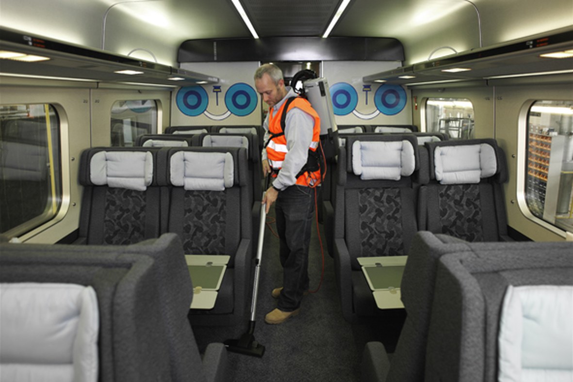 Operator with a Nilfisk GD5 backpack vacuum cleaning between fabric seats inside a passenger train carriage during turnaround