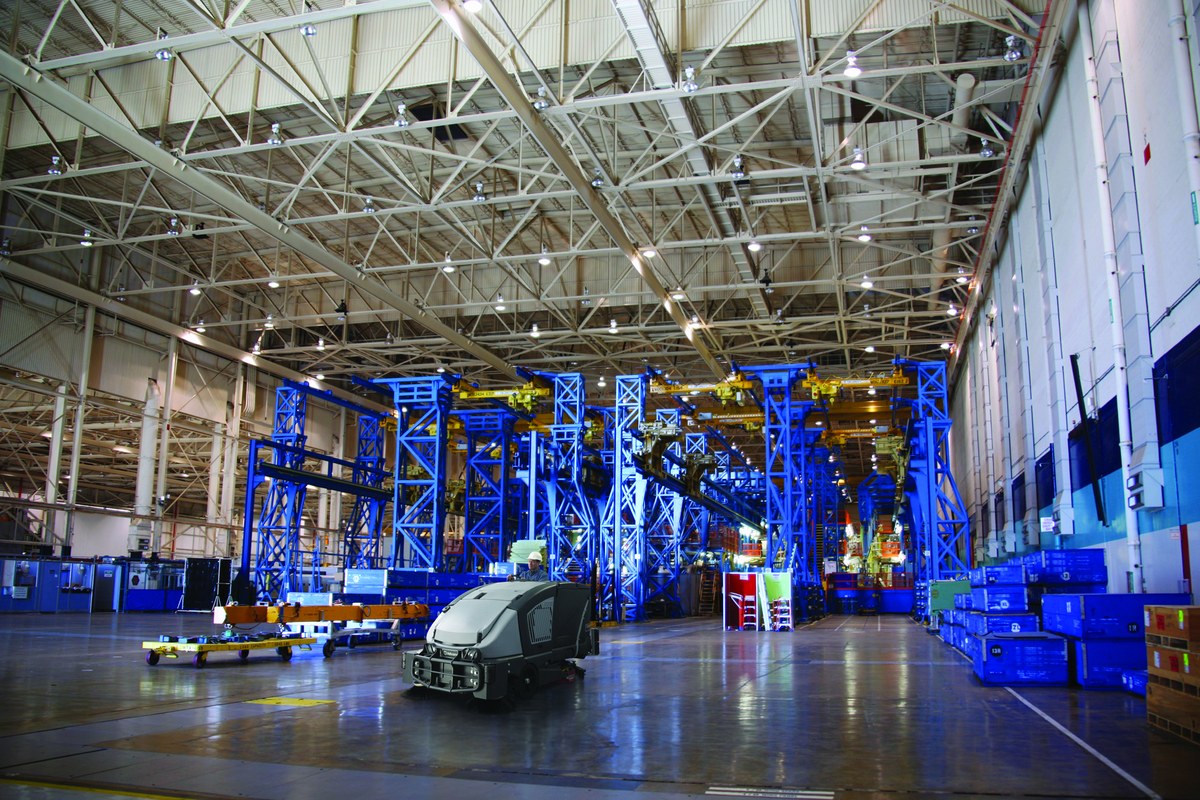 Operator on a Nilfisk CS7000 combination sweeper-scrubber cleaning the polished concrete floor of a large aircraft hangar with industrial gantry framework overhead