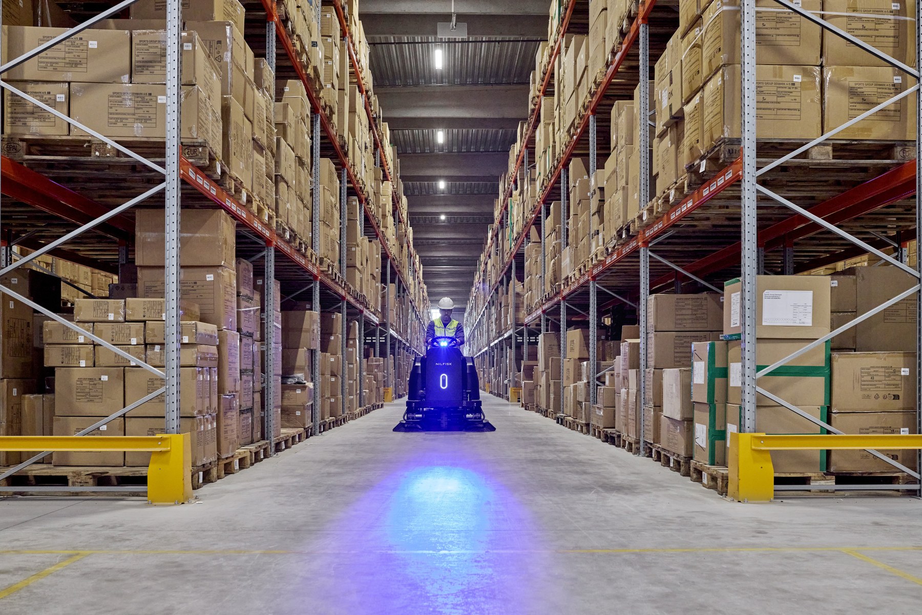 Operator on a Nilfisk ride-on scrubber dryer with UV-C disinfection lamp cleaning a tall pallet-racking aisle in a large distribution warehouse