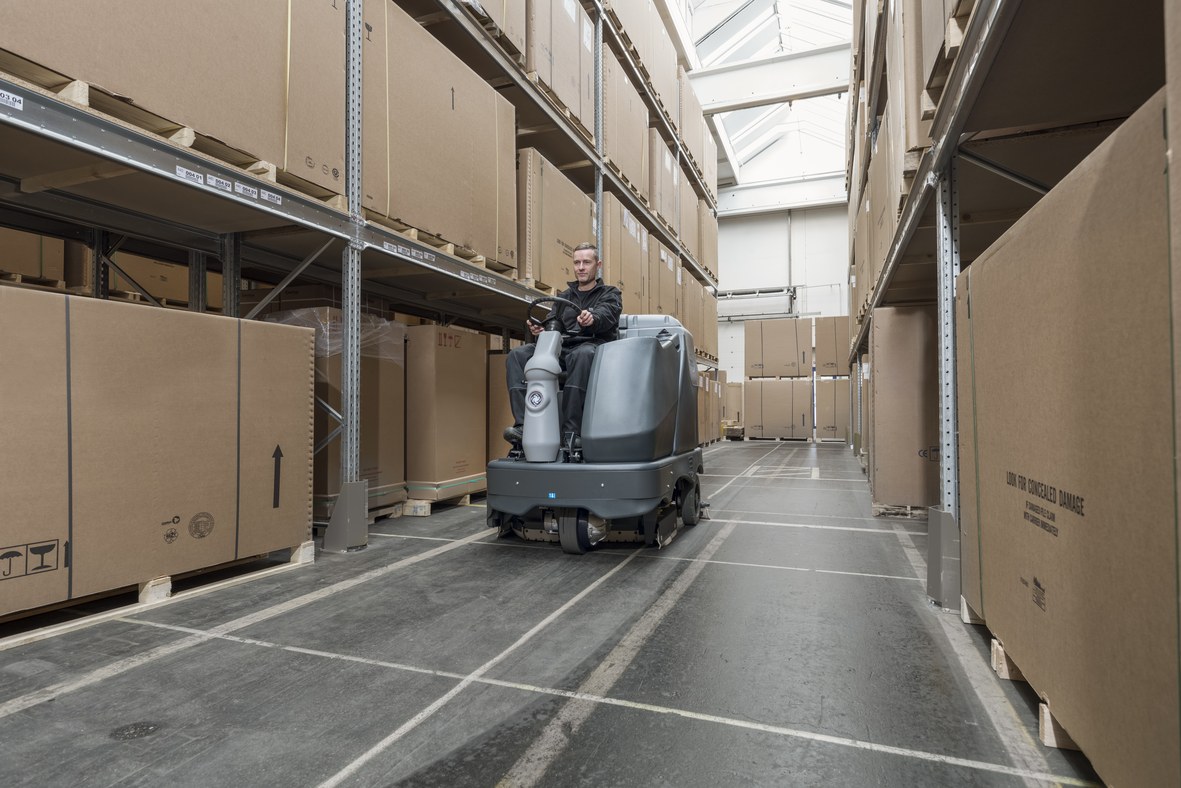 Operator on a compact Nilfisk ride-on scrubber dryer cleaning between cardboard cartons stacked on pallet racking in a warehouse aisle