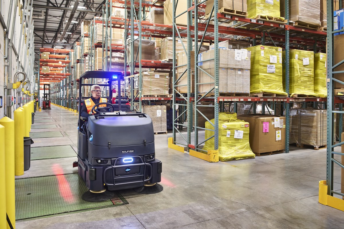 Operator on a Nilfisk CS7500 combination sweeper-scrubber cleaning a busy industrial warehouse aisle lined with stretch-wrapped pallets